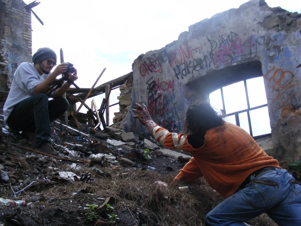 A person with long hair and an orange shirt reaches out towards a man taking a photograph among the ruins of a burned building, with graffiti on the wall.