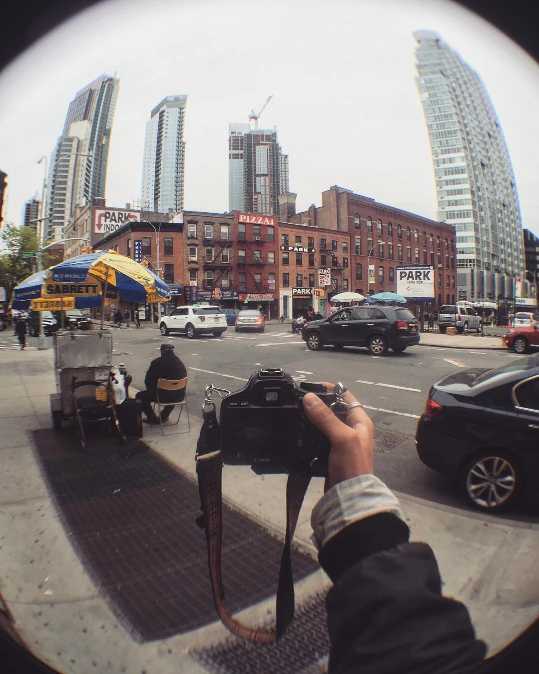 A street scene with a person taking a photo using a camera, parked cars, a man sitting on the sidewalk, and tall buildings in the background.