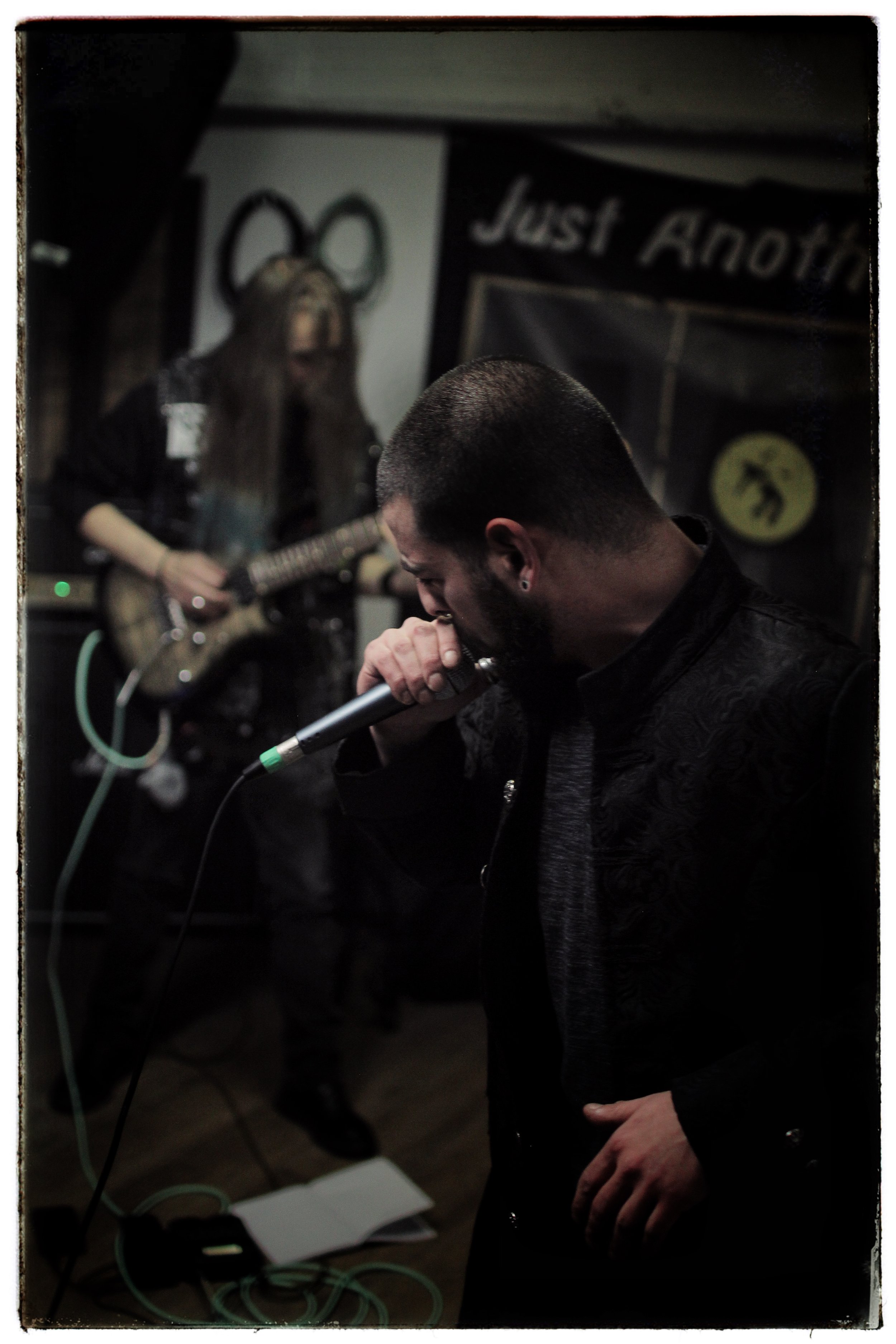 A man with short hair and a beard is singing into a microphone, and in the background, another person with long hair playing an electric guitar. The setting appears to be a small, dimly lit music venue with a banner that reads 'Just Another' on the w