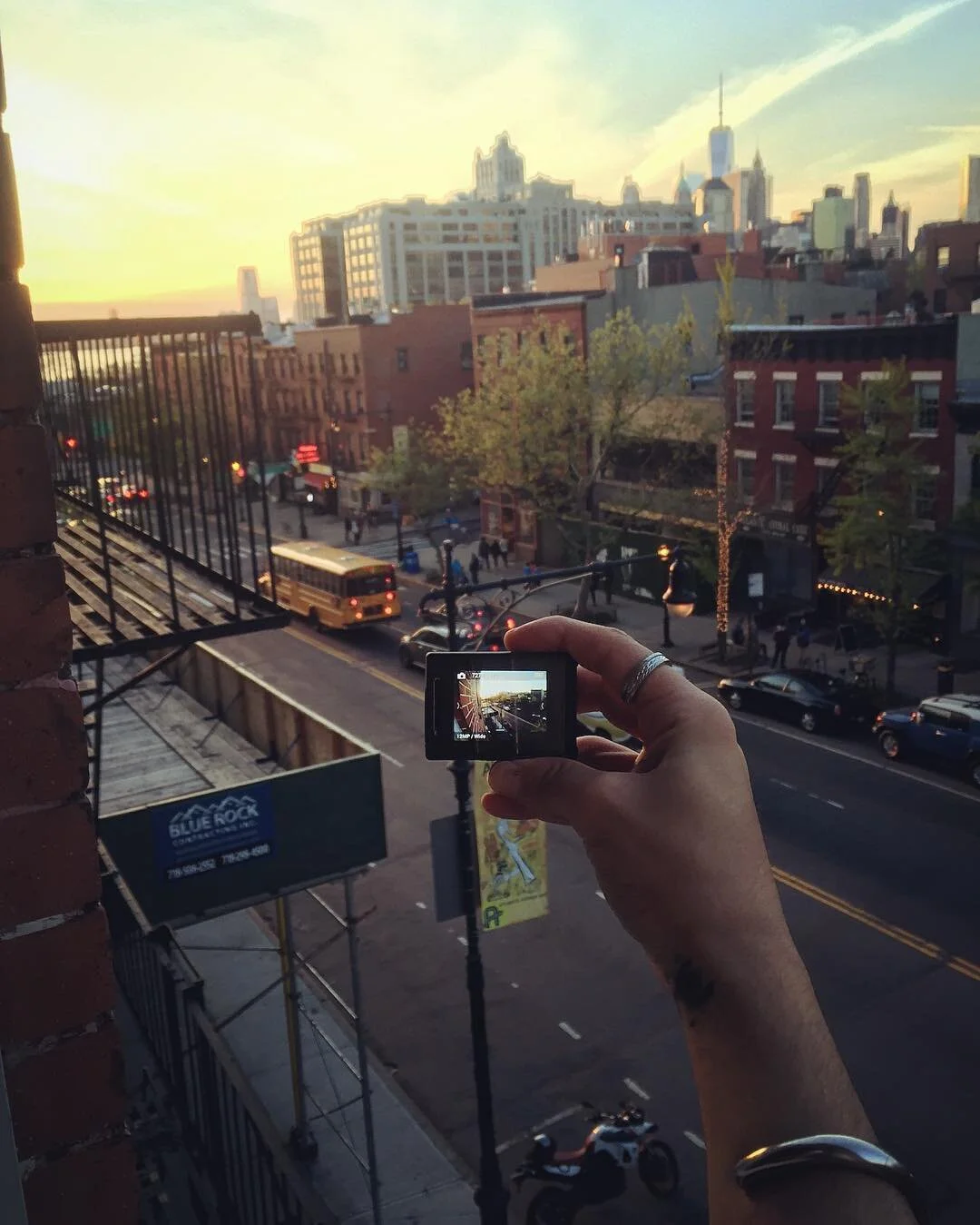 A hand holding a small camera taking a picture of a city street scene at sunset, with buildings, trees, a bus, cars, and pedestrians visible.