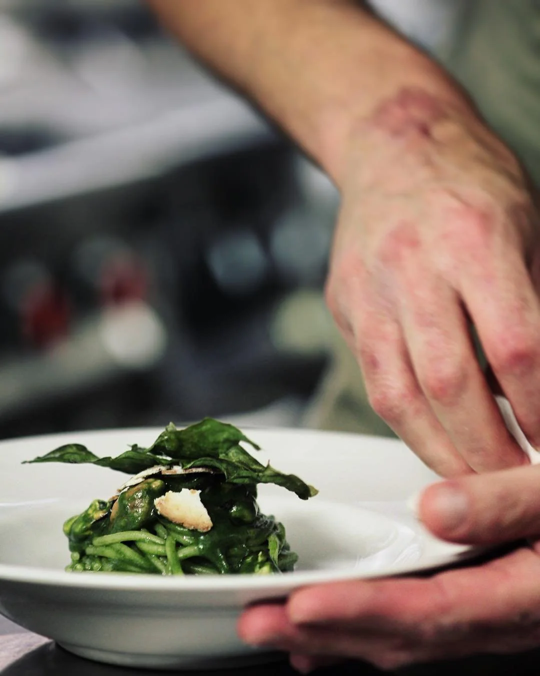 A person's hand garnishing a plate of pasta with greens in a professional kitchen setting.