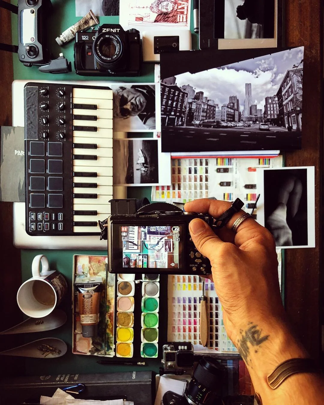 A photographer's workspace with various creative tools including a camera, a keyboard, paint watercolors, printed photographs, a miniature camera, and a laptop, all arranged on a desk.