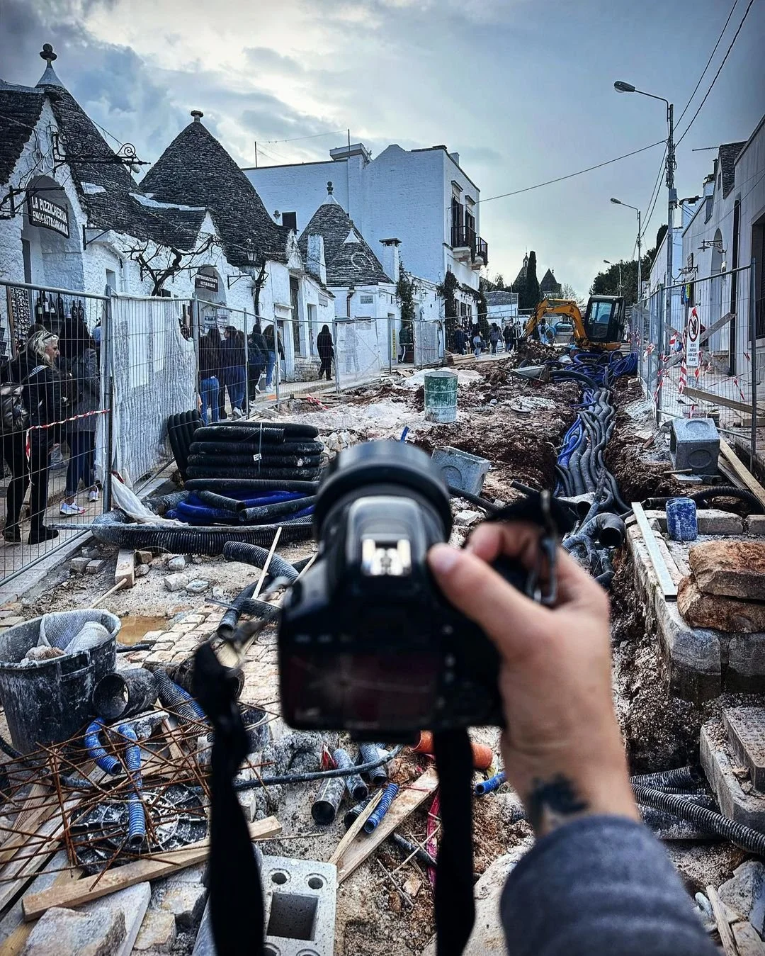 Street under construction with a person holding a camera in the foreground, construction equipment, pipes, and a group of people observing behind a fence.