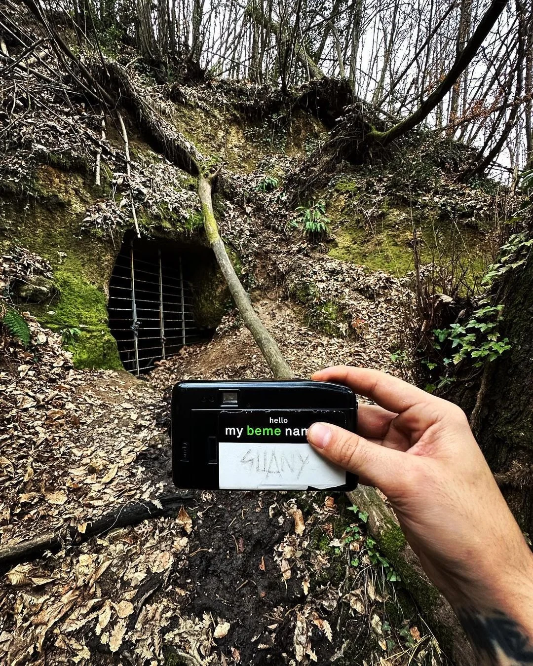 Person holding a camera with a note saying 'Hello my beme name' in front of a mossy wooded area with a small barred opening in a hillside.