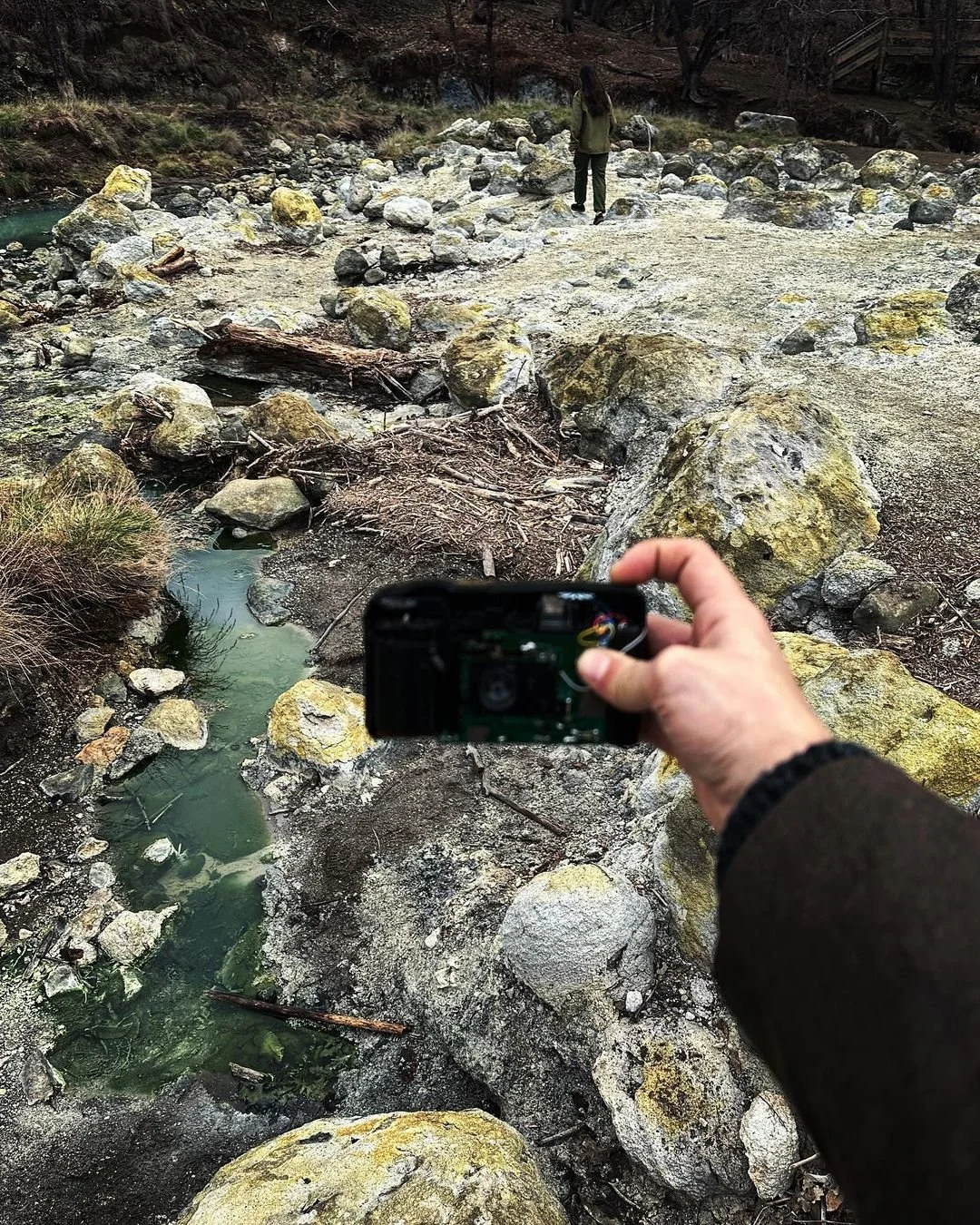 Person holding a camera taking a photo of a person walking on a rocky landscape with small water pools and scattered rocks and logs, background of leafless trees.
