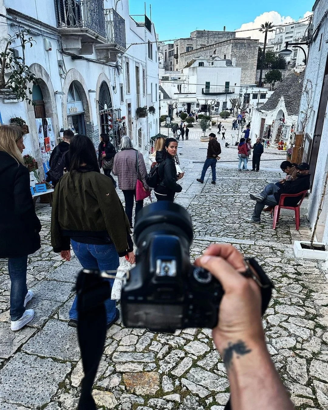 A cobblestone street in a Mediterranean town with white buildings and small shops, bustling with people, some sitting on benches, others walking around, under a blue sky with a few clouds.