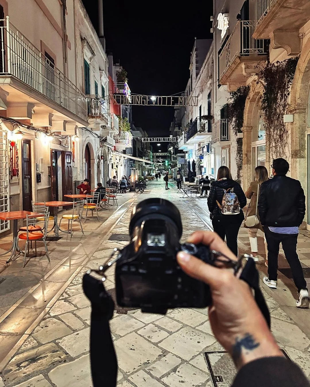Nighttime street scene with people walking, outdoor tables, and buildings with balconies. A person is holding a camera, taking a photo of the street.