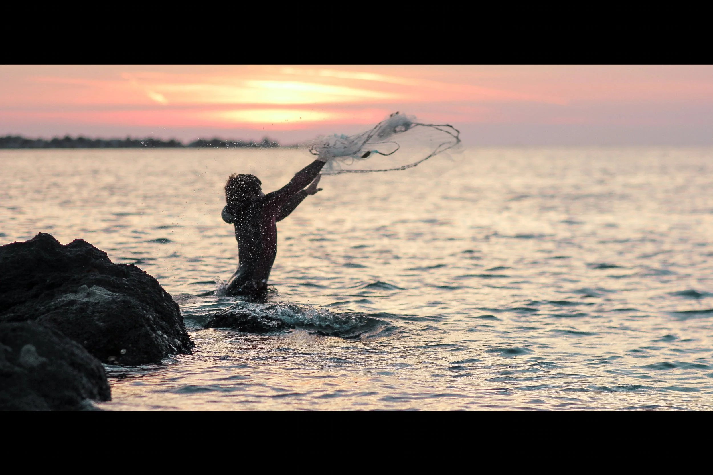 Person standing on rocks at the edge of the water, throwing a net during sunset