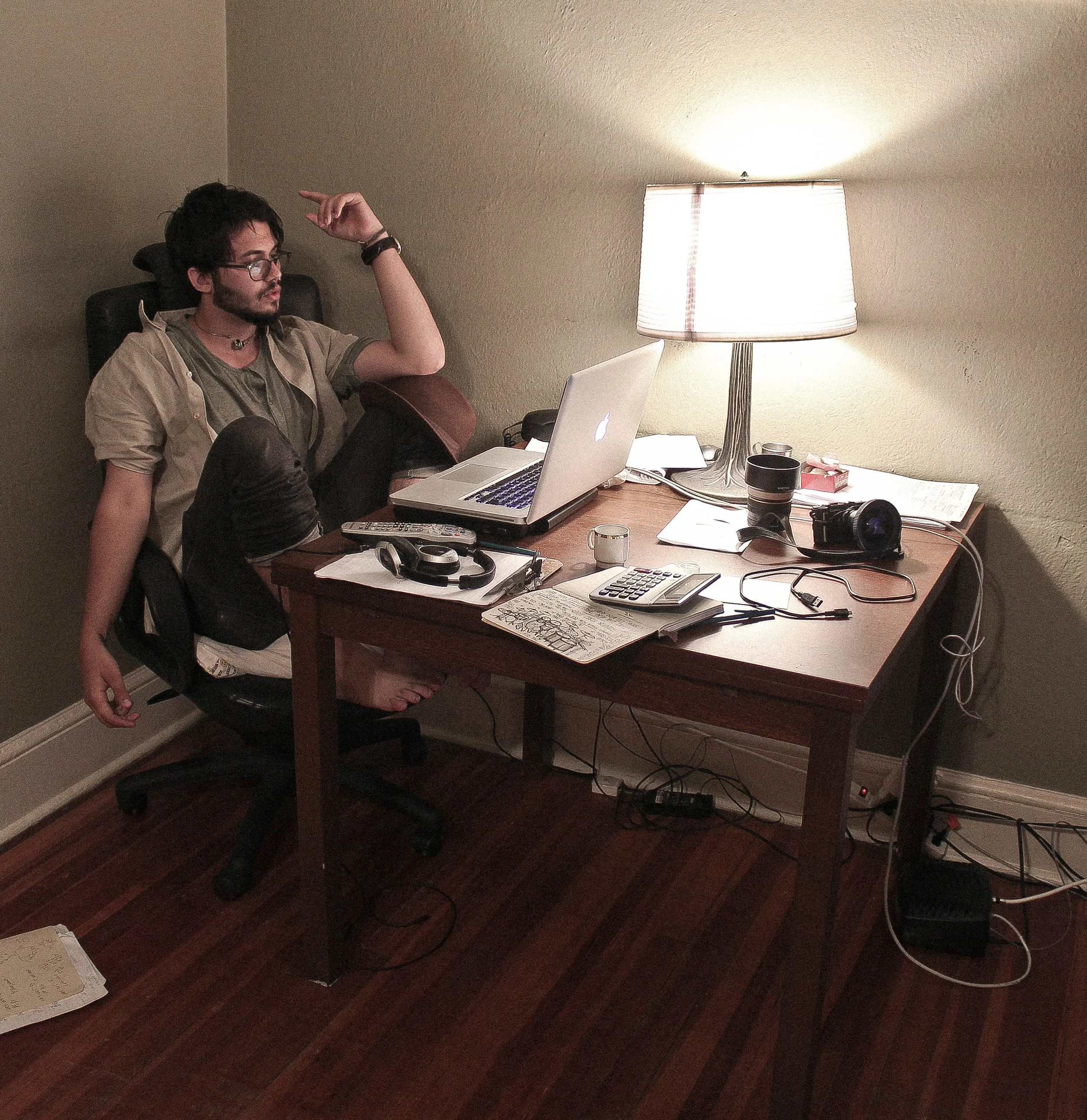 A person with dark hair, glasses, and a beard sitting in a chair at a cluttered desk, with a lamp casting light on the wall behind him, using a laptop.