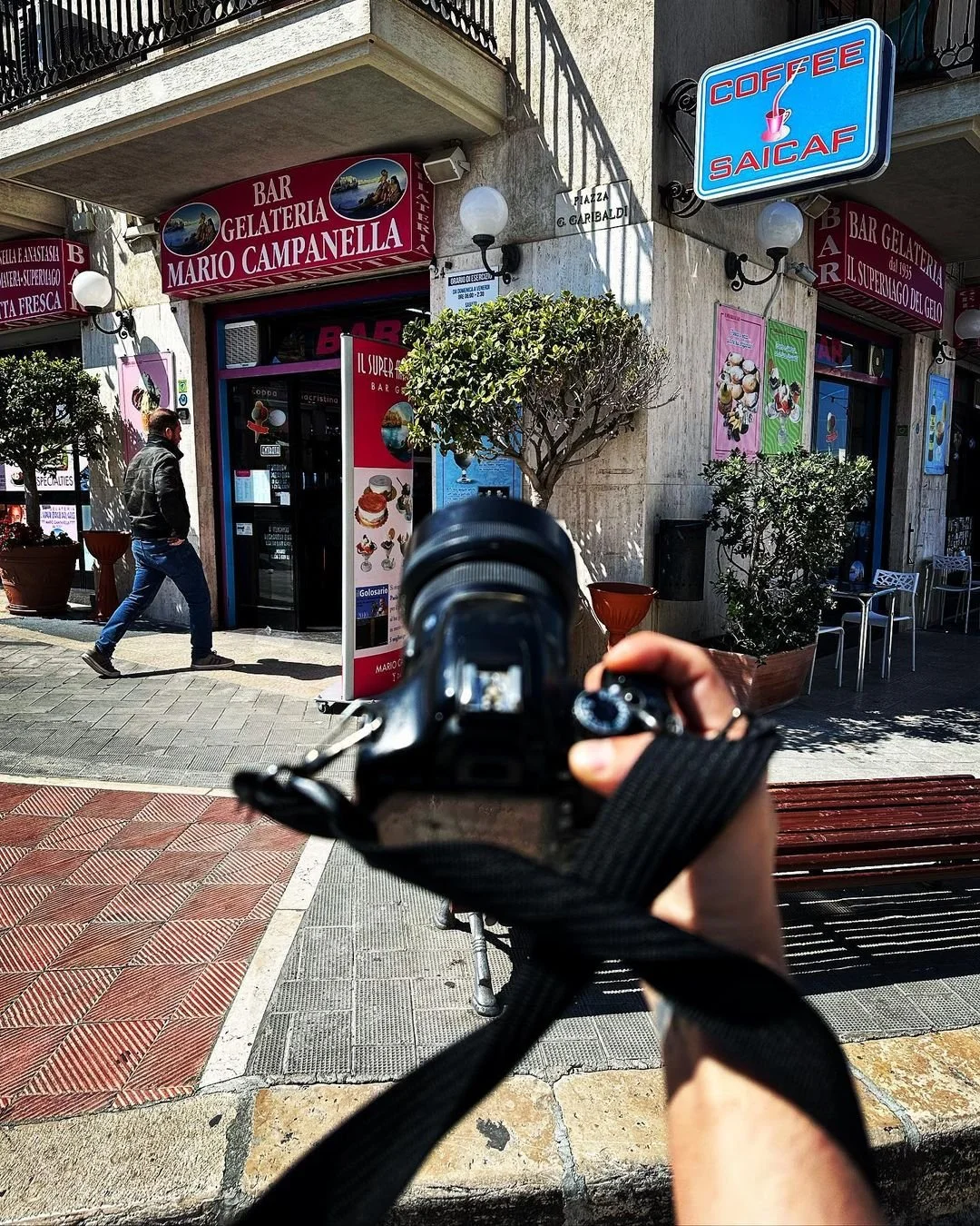 A person holding a camera, taking a photo of a gelato and coffee shop on a city street. The shop has pink and purple signage, trees in planters, and a man walking past.