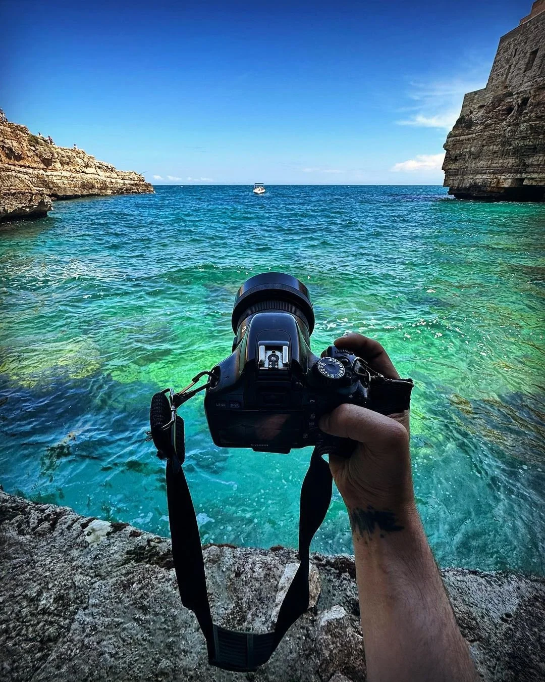 A person holding a camera captures a view of turquoise water between rocky cliffs with a boat in the distance under a clear blue sky.