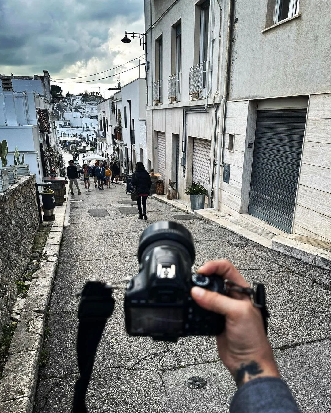 A person holding a camera takes a photo of a narrow street with pedestrians and white buildings, under cloudy skies.