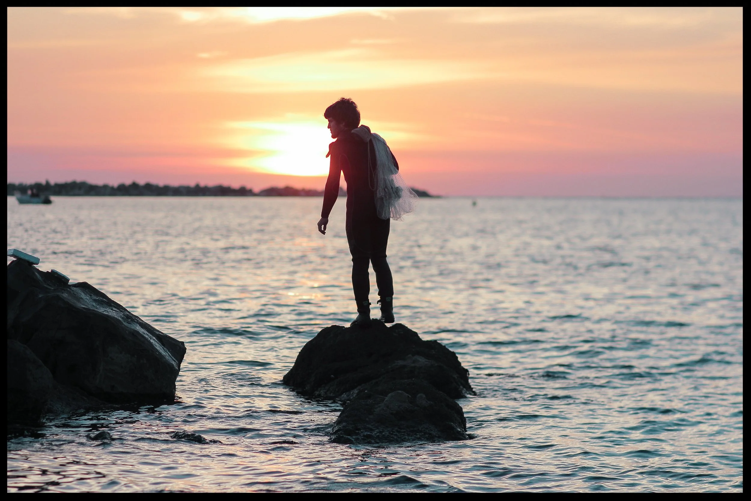 A person standing on rocks by the water during sunset, with a backpack and fishing net, gazing at the horizon.