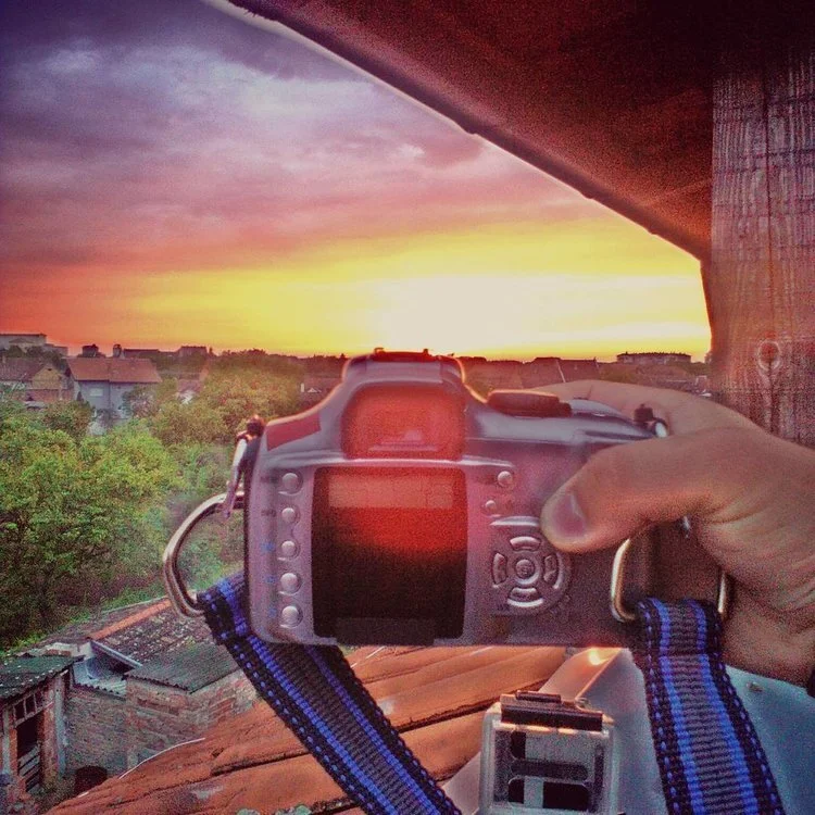 A digital camera is being held up by a person's hand, with a sunset sky featuring pink, purple, and yellow hues in the background. The scene is outdoors, with trees and rooftops visible below.