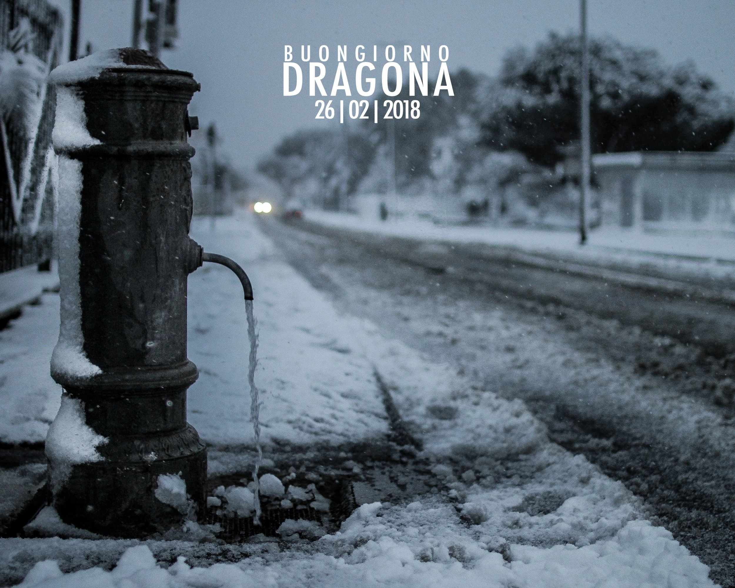 Close-up of an old fire hydrant covered in snow, with a snowy street and trees in the background, and a message greeting 'Buongiorno Dragona' along with the date '26 | 02 | 2018' overlaid on the image.