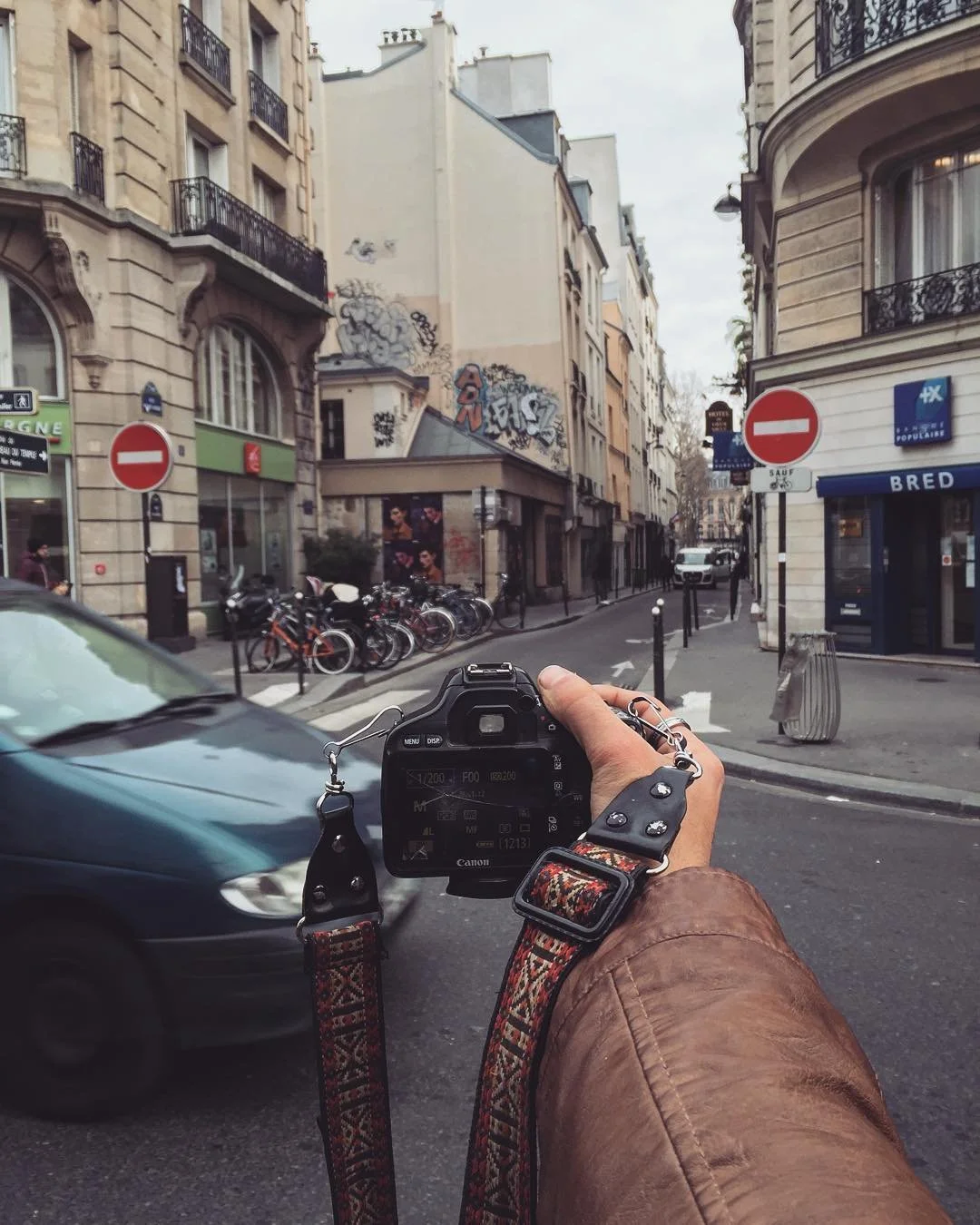A person holding a camera on a city street, with buildings, parked bicycles, and moving cars, and street signs visible.