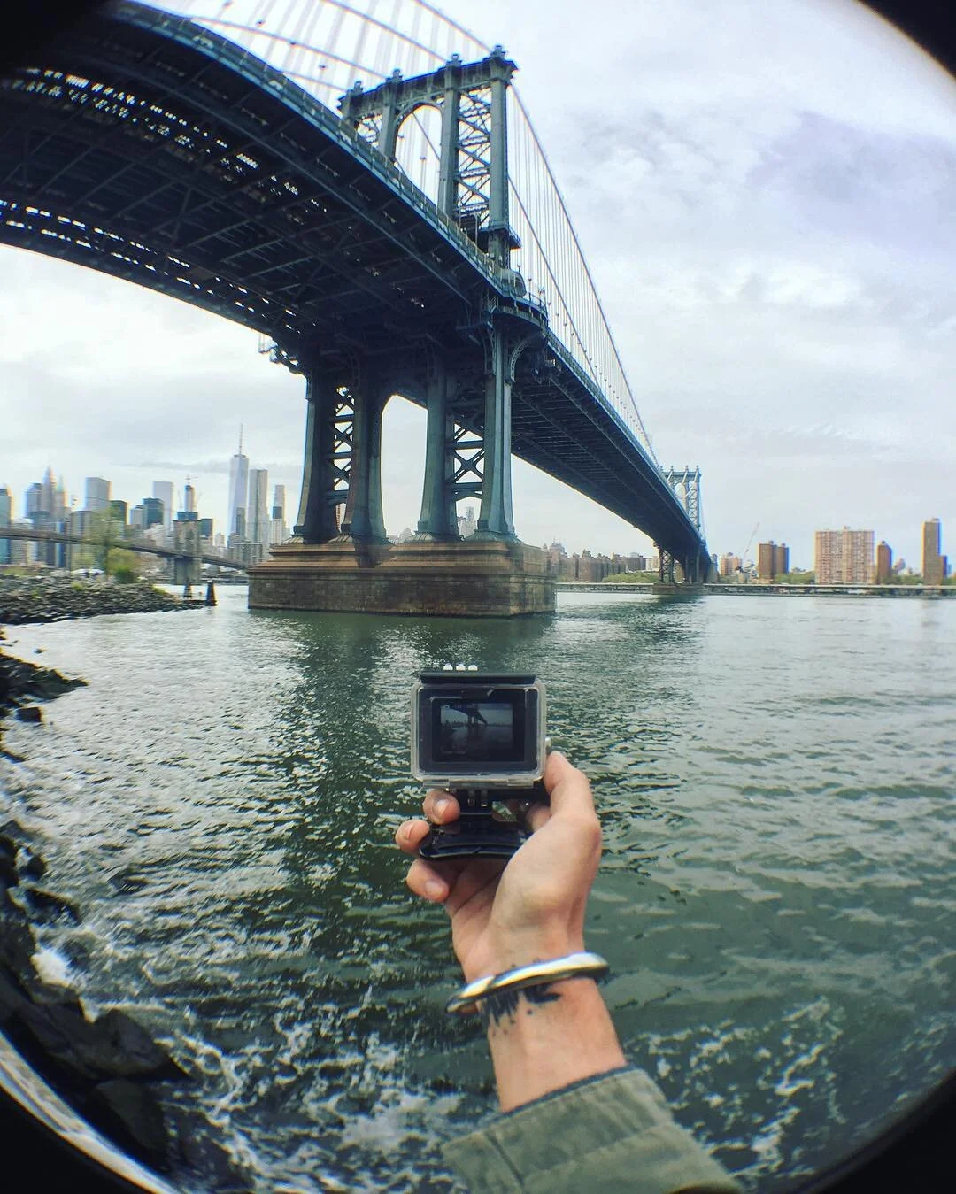 View of a large suspension bridge over water with a city skyline in the background, taken with a GoPro camera held by a person.