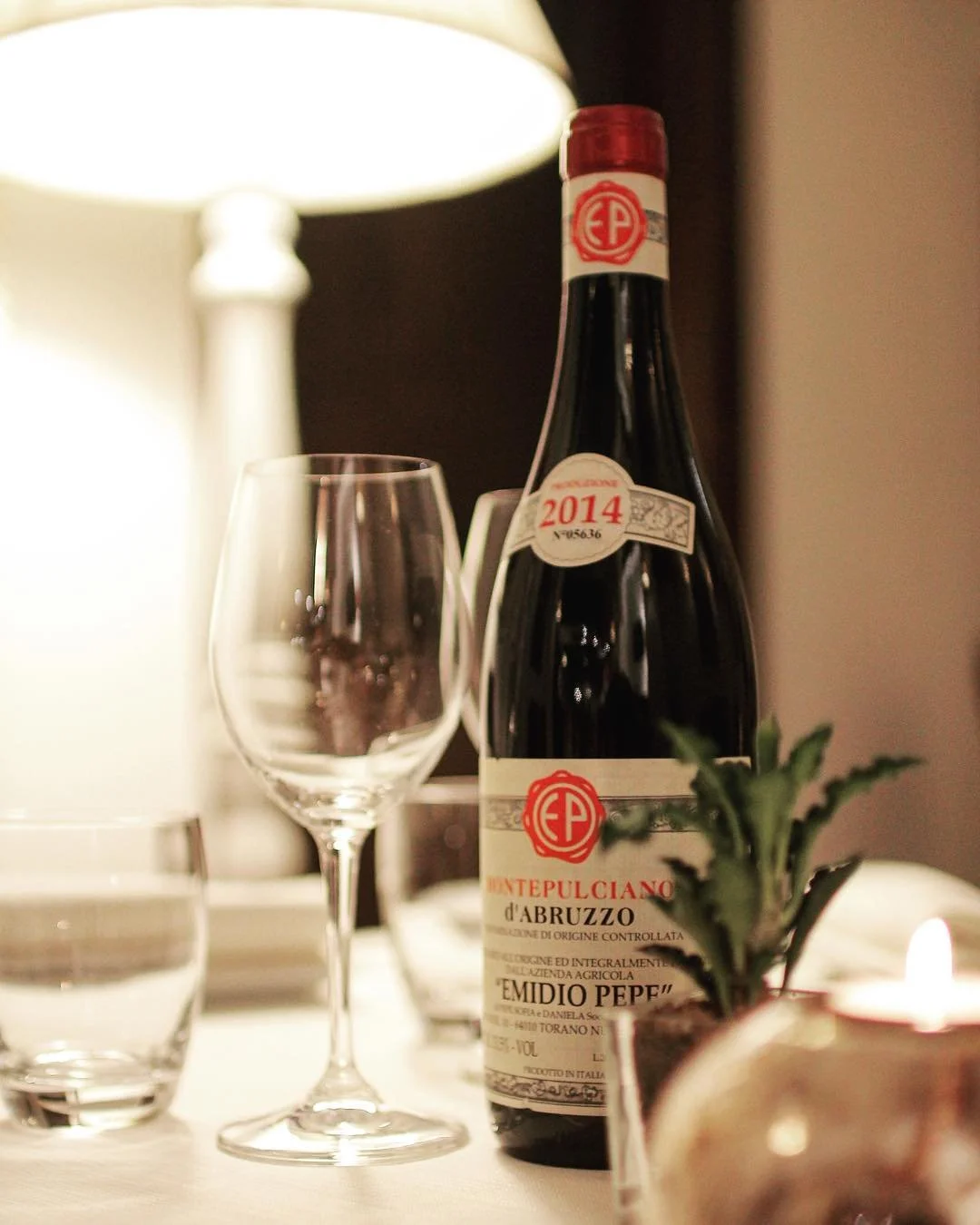 A bottle of red wine labeled 'Montepulciano d'Abruzzo' with vintage year 2014, a wine glass, a lit candle, a small potted plant with green leaves, and a lamp in the background on a table.