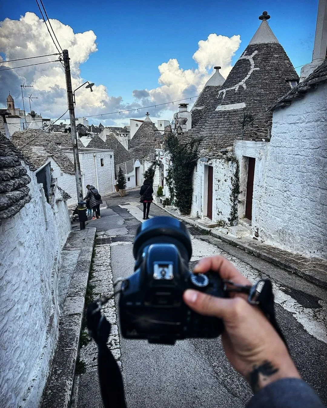Person holding a camera on a street with white-washed buildings and cone-shaped roofs, some with exterior ivy, in a scenic village with cloudy sky.