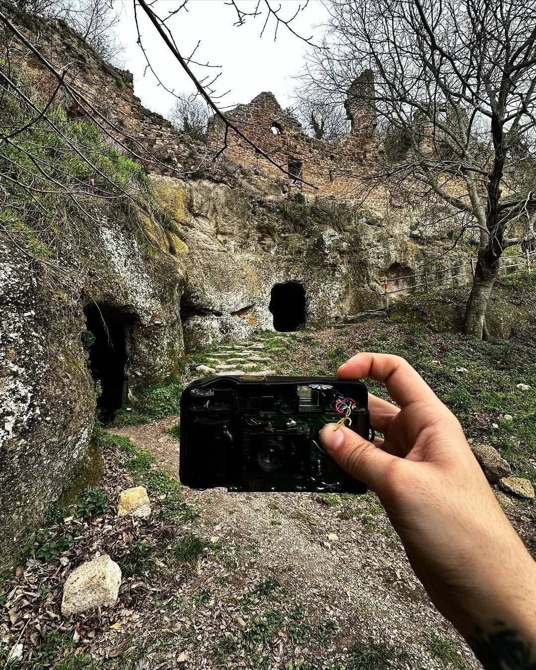 A hand holding a disassembled camera in front of an ancient rock cave with two dark openings, set in a rocky and wooded landscape with leafless trees.