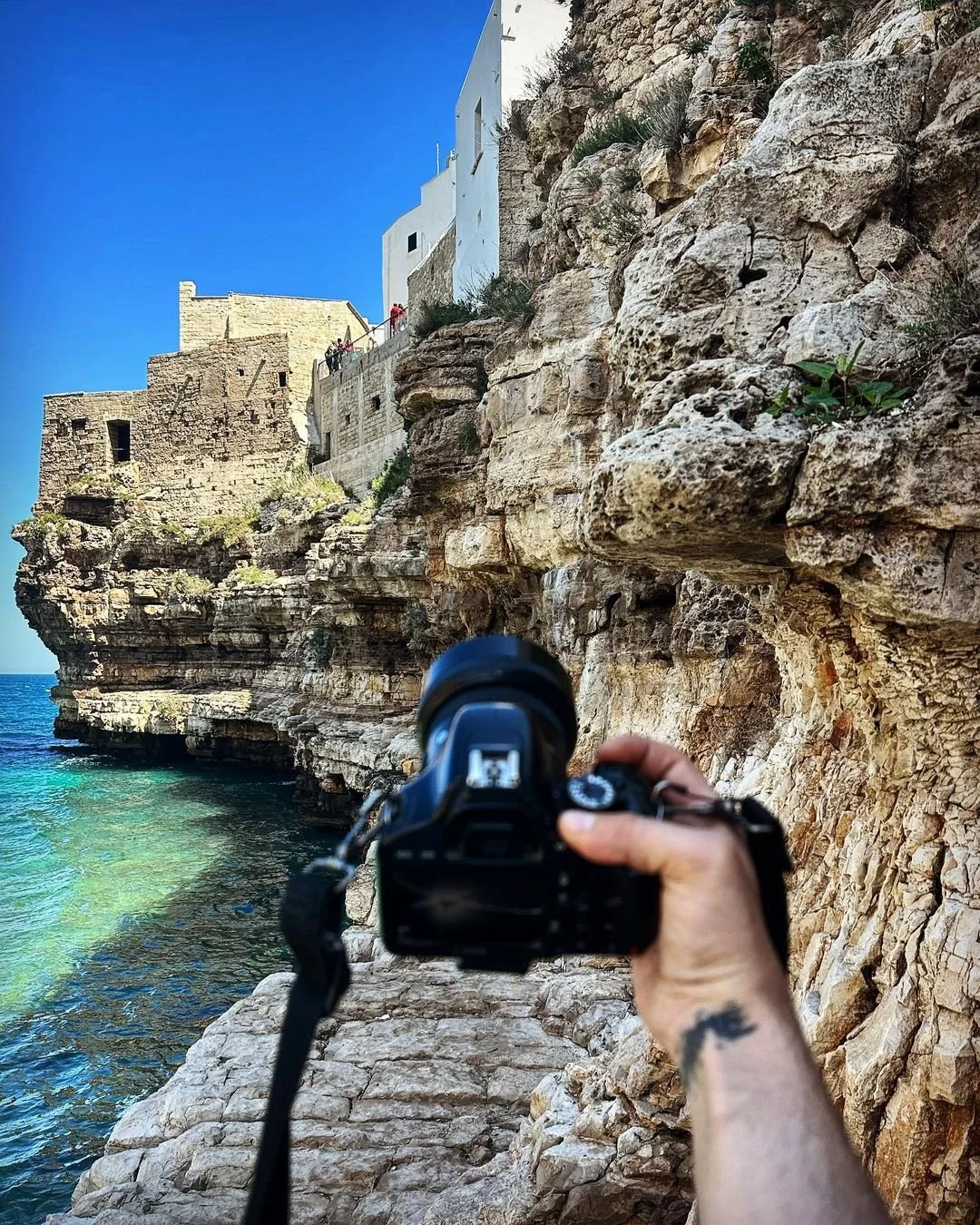 Person taking a photograph along a rocky coast with water in the foreground and buildings on a cliff above.
