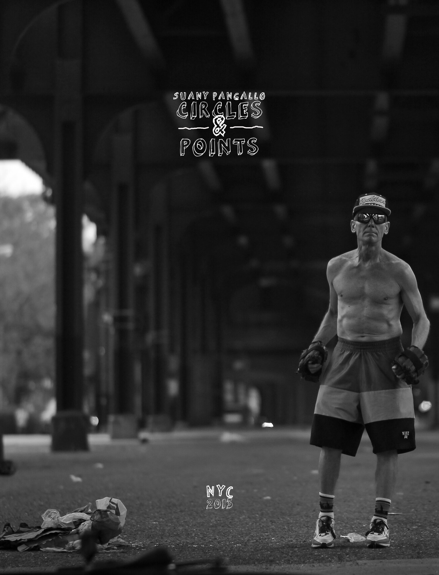 A shirtless man wearing shorts, sneakers, sunglasses, and a cap stands under a bridge in NYC in 2013. There is trash on the ground in front of him.