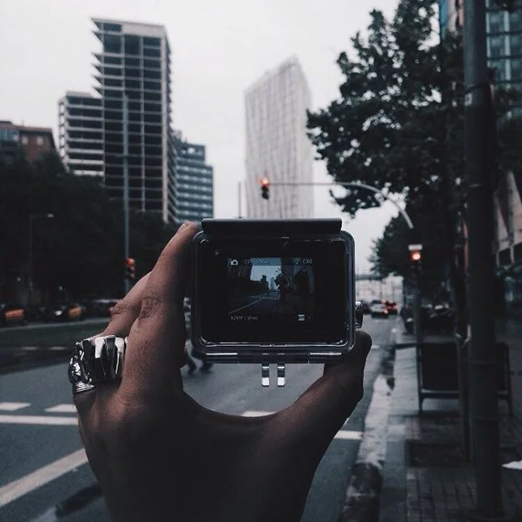 A person holds a camera capturing a city street scene with tall buildings and traffic lights, on an overcast day.