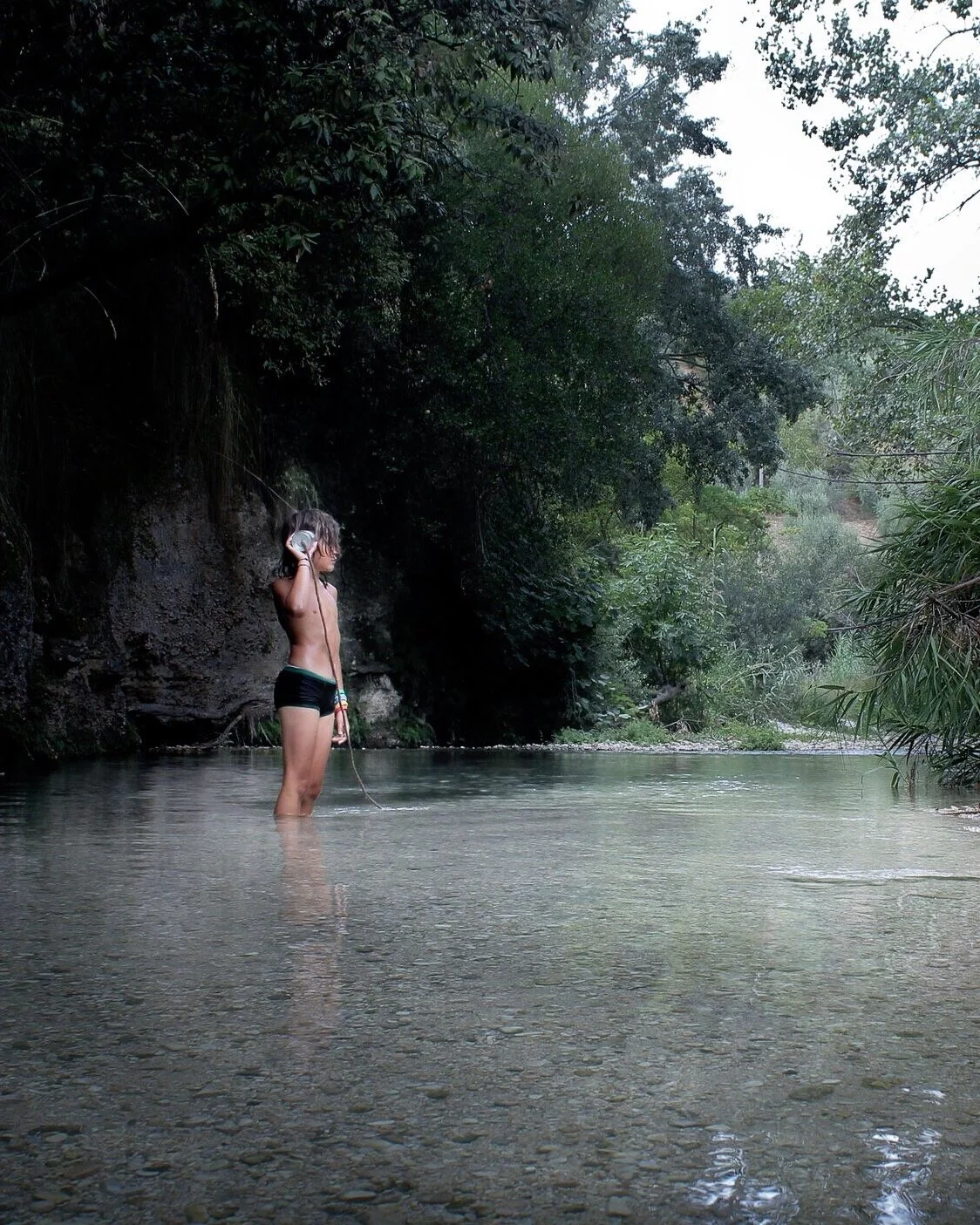 A young girl standing in a shallow river, holding a phone to her ear, surrounded by lush green trees.