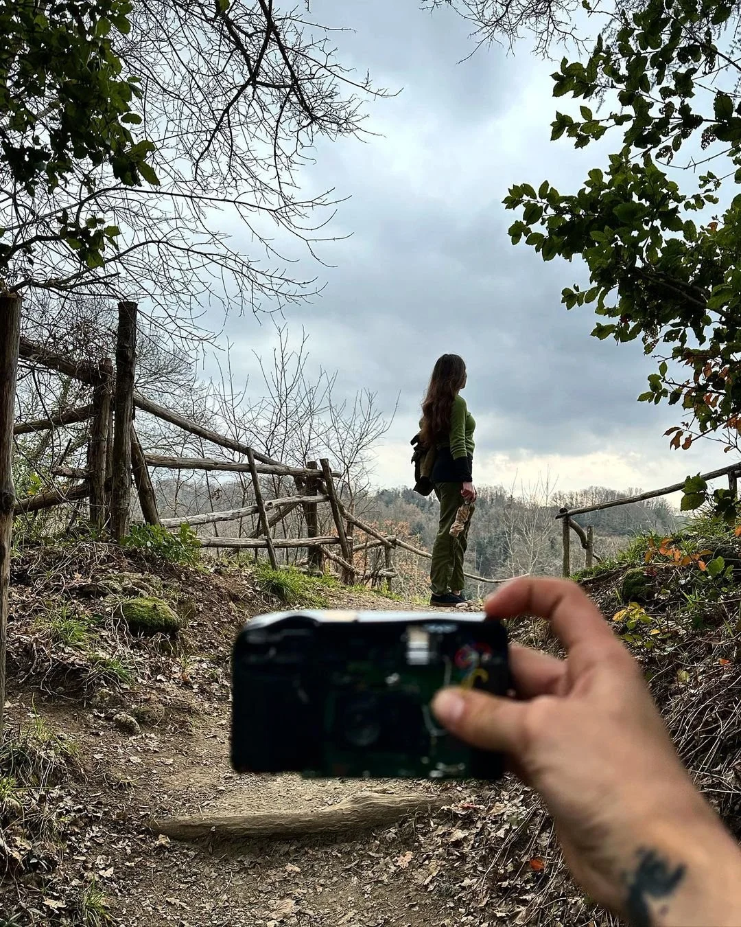A woman standing on a hiking trail, being photographed with a camera by a person with a visible tattoo on their hand. The trail has wooden railings on both sides, trees with sparse leaves, and an overcast sky.