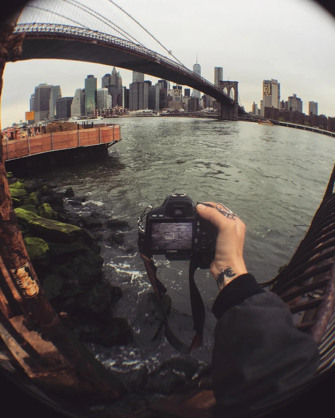 A person holding a Canon camera captures a view of the Brooklyn Bridge and Manhattan skyline from the water in New York City.