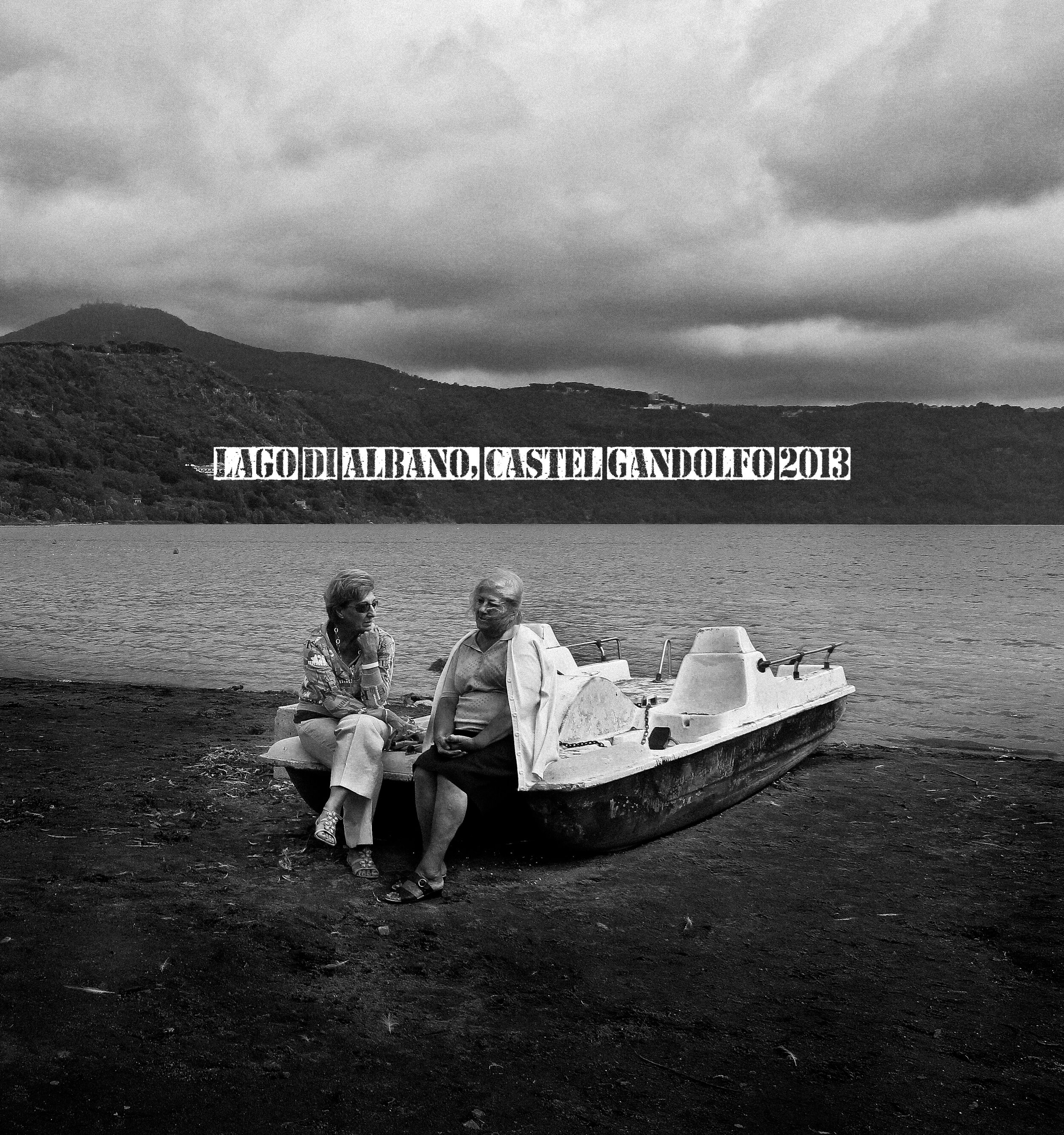 Black-and-white photo of two older adults sitting on a boat on Lake Albano in Castel Gandolfo, Italy, with hills and cloudy sky in the background.