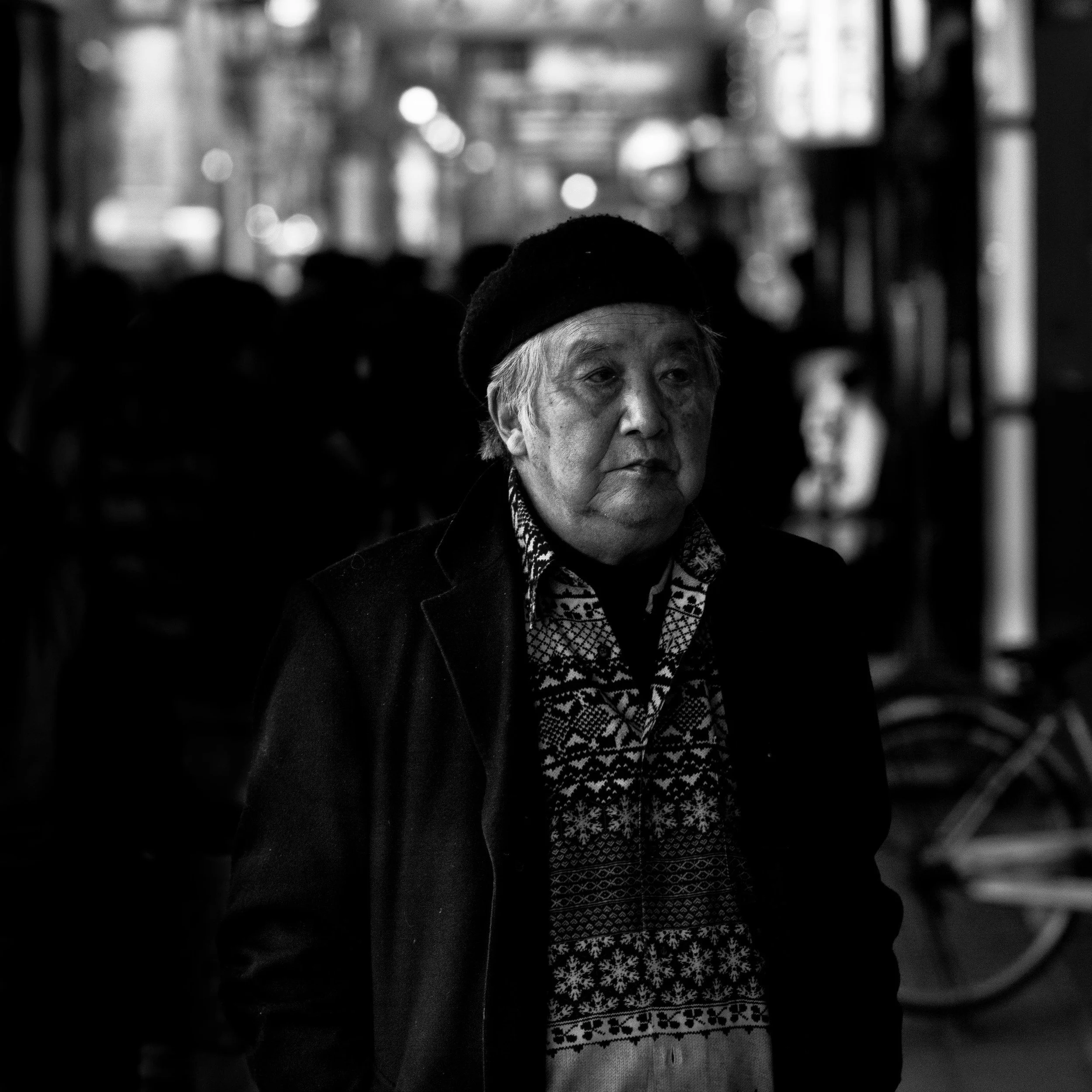A black and white photo of an elderly man with gray hair, wearing a knit hat, patterned scarf, and patterned shirt, in a crowd at an indoor event or gathering.