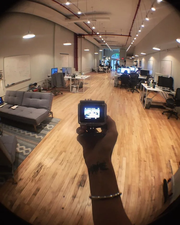 Open office space with multiple desks and computers, a gray couch with a rug, and a hand holding a camera in the foreground capturing the room.