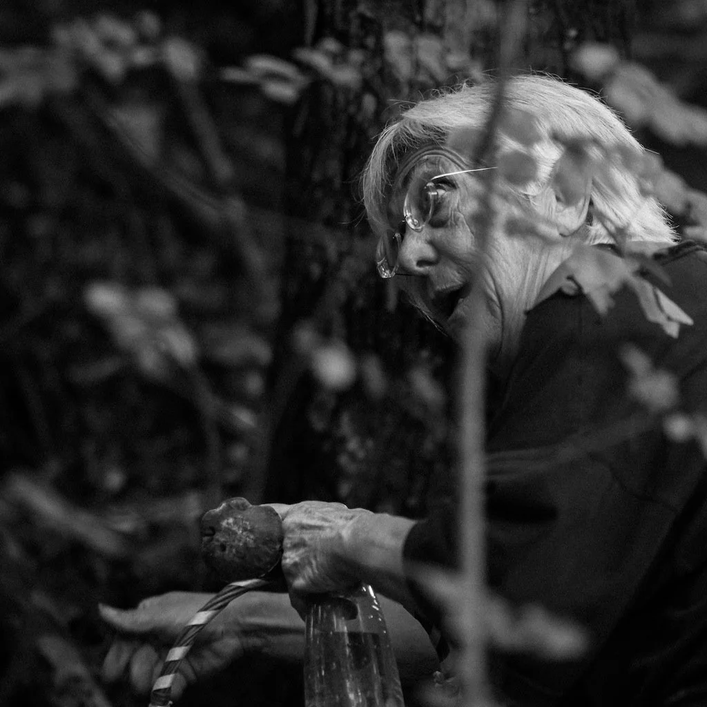 An elderly woman with glasses is smiling and tending to plants or a garden, with a large pile of logs or wood in the background.