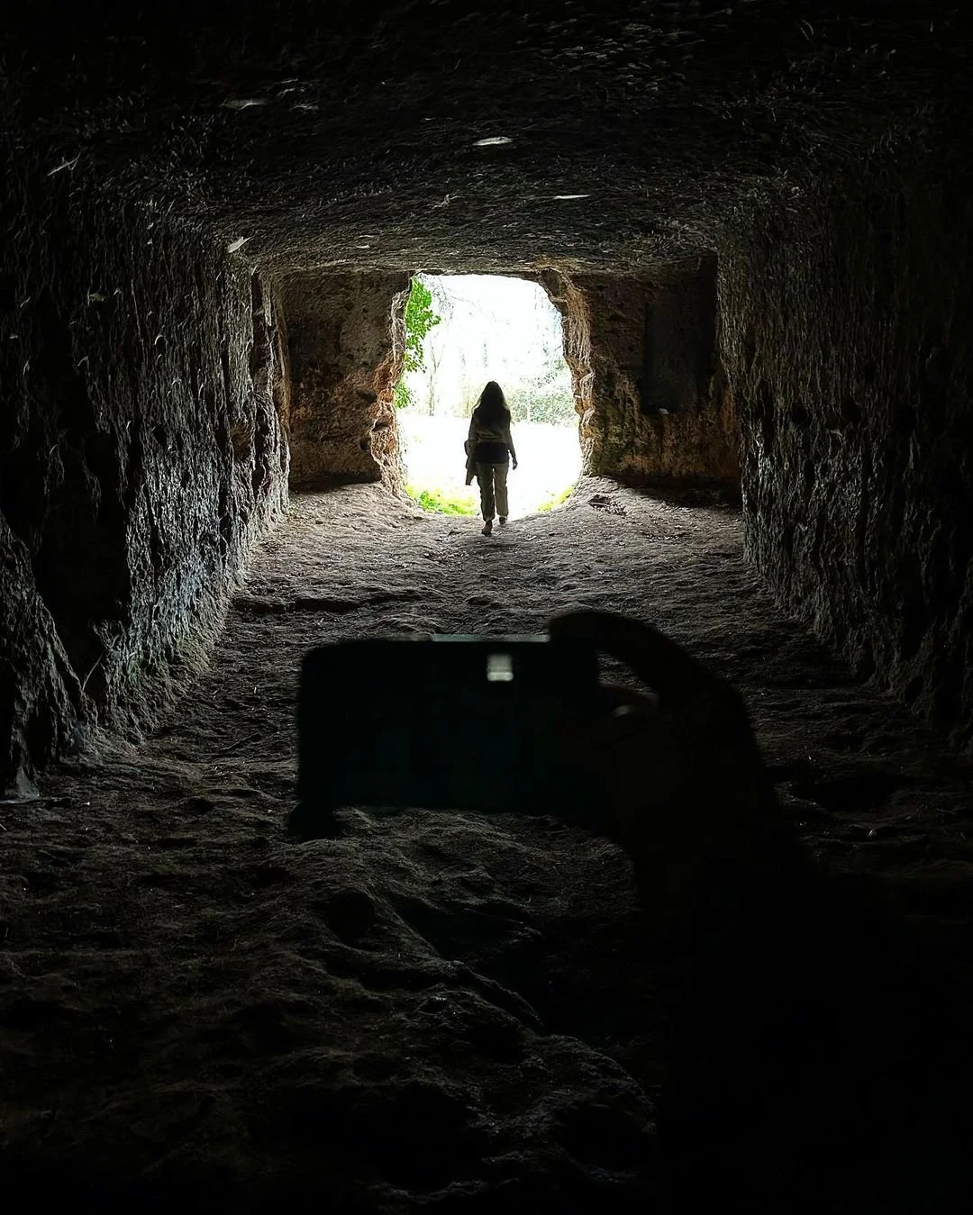 A person walking out of a dark cave towards a bright outdoor area, with a camera capturing the scene from inside the cave.