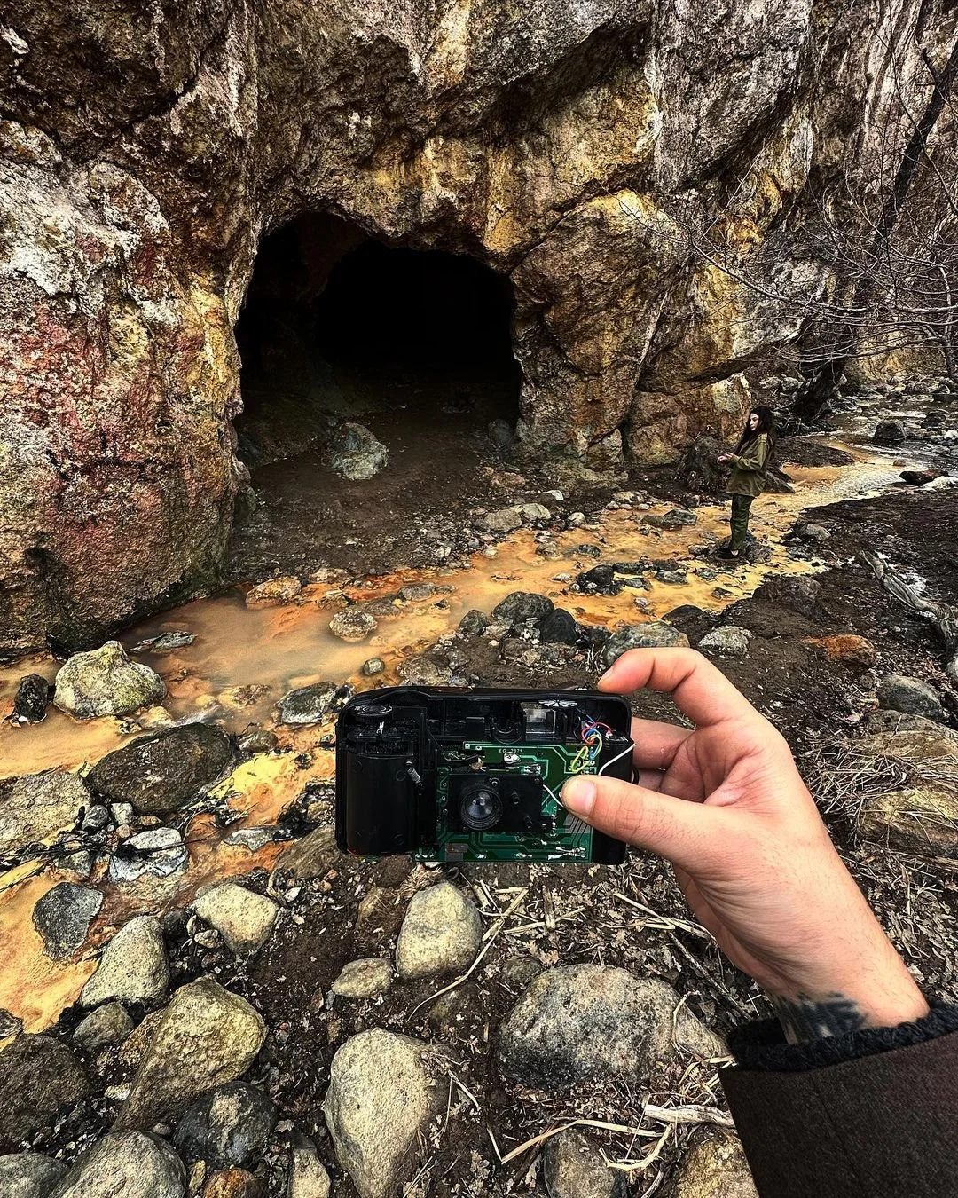 A person holding a partially disassembled camera in front of a rocky creek in a wooded area. There is a woman standing in the background near the creek, facing a dark cave or tunnel in a large rock formation.