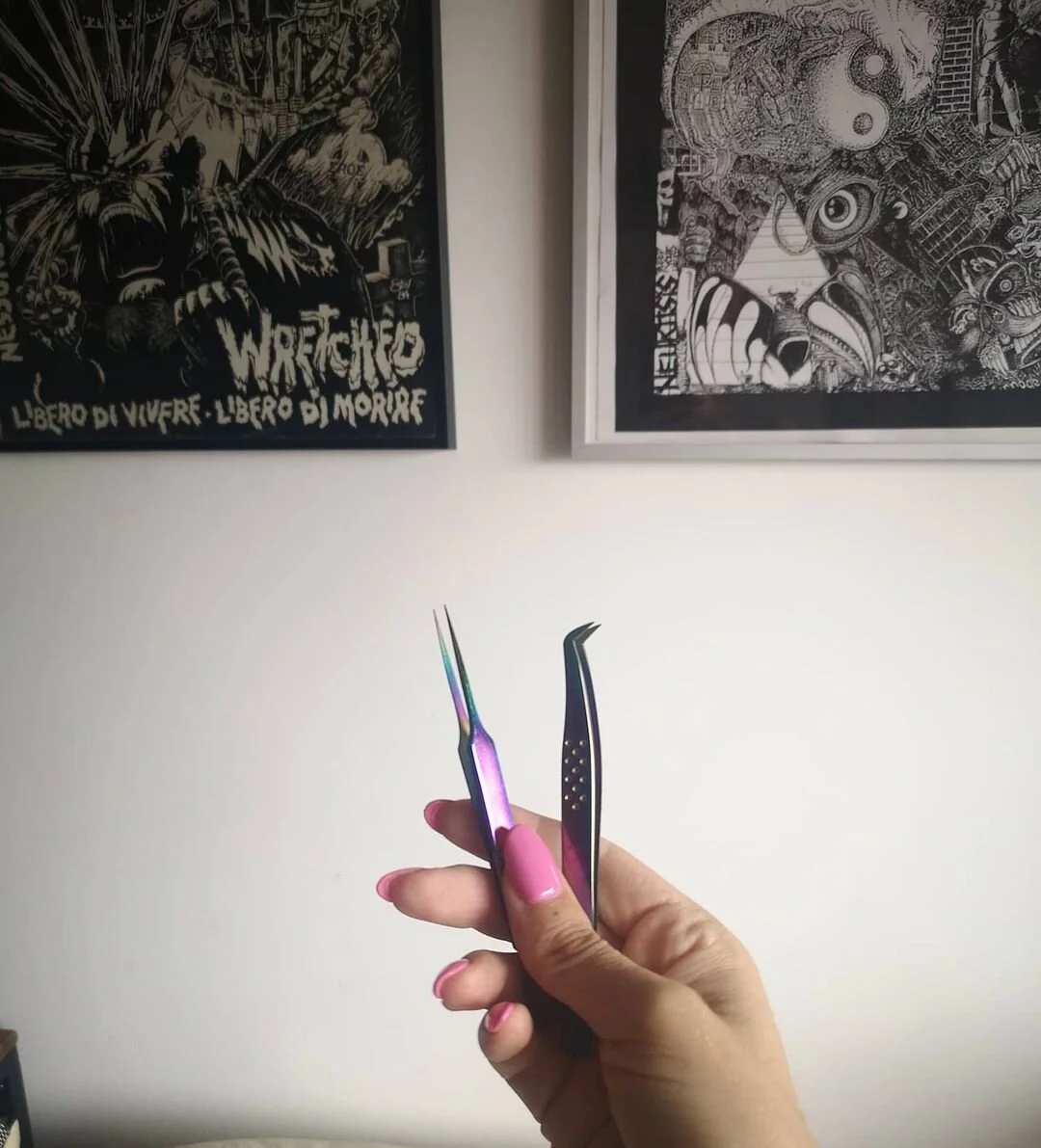 Person holding two black and iridescent tweezers with pink nails, two art posters on the wall, one with a wolf face and the words 'Liberi di vivere. Libero di morire,' and the other with an abstract face and various designs.