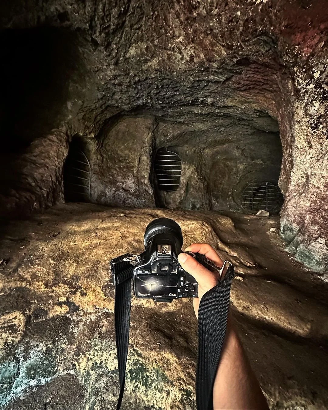 A person holding a camera inside a dark natural cave, with three metal grate coverings on the back wall of the cave.