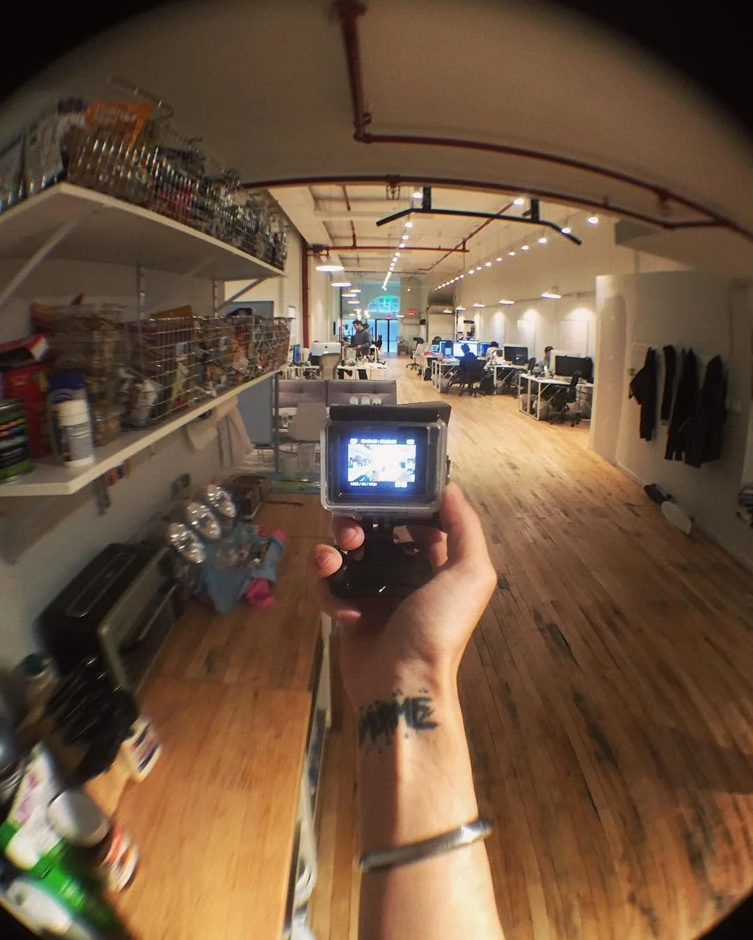 Interior of an office space with desks, office chairs, and computers, photographed through a fisheye lens with a person holding a camera in the foreground.