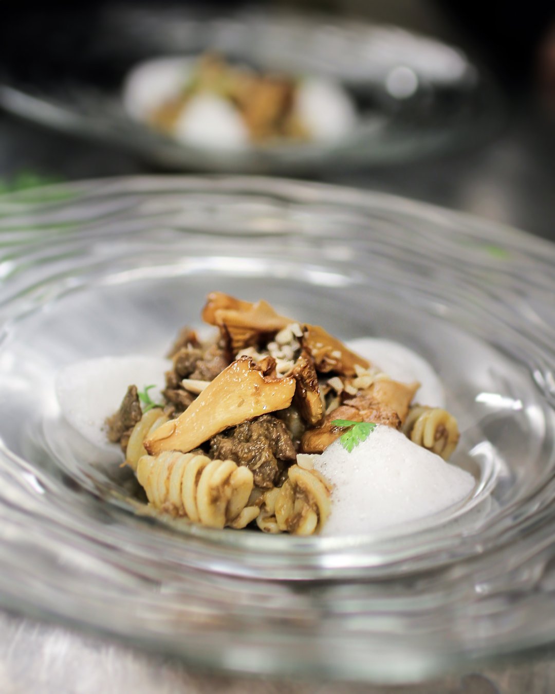 Close-up of cooked pasta with mushrooms and ground meat garnished with a small parsley leaf, served with foam in a glass dish.