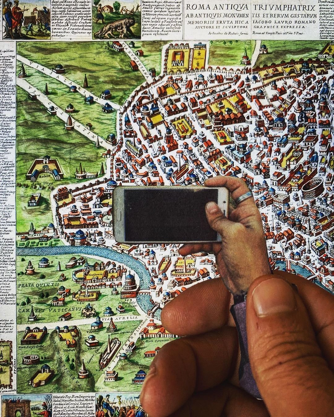 A person holding a smartphone taking a picture of a colorful historical map of Rome, Italy, showing streets, buildings, and landmarks.