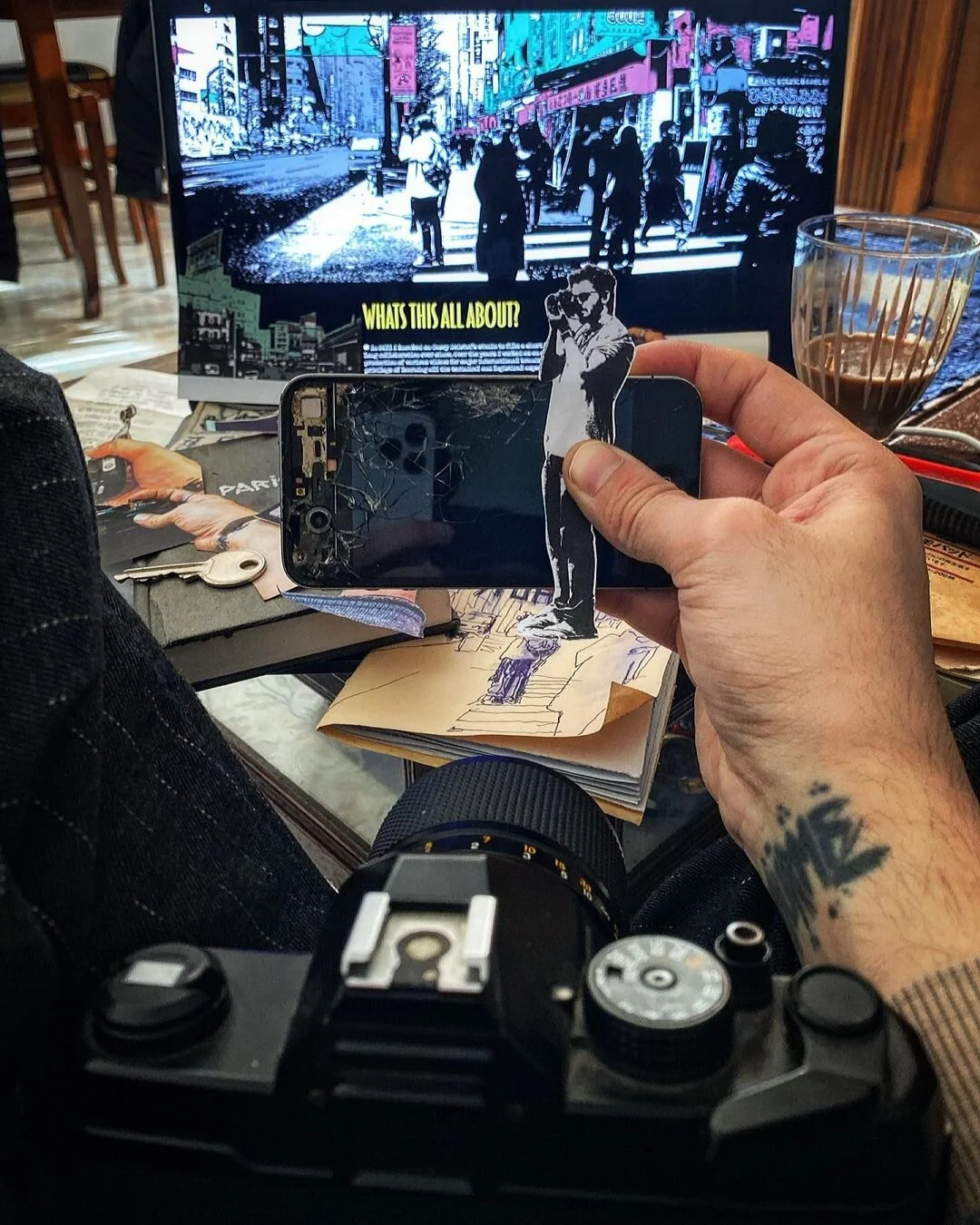 A person holding a cracked smartphone with a sticker of a person taking a photo in front of a computer screen. The scene is cluttered with a camera, sketchbook, keys, and a glass of coffee. The computer screen displays a stylized image of a city street with pedestrians and a caption that reads "WHAT'S THIS ALL ABOUT."