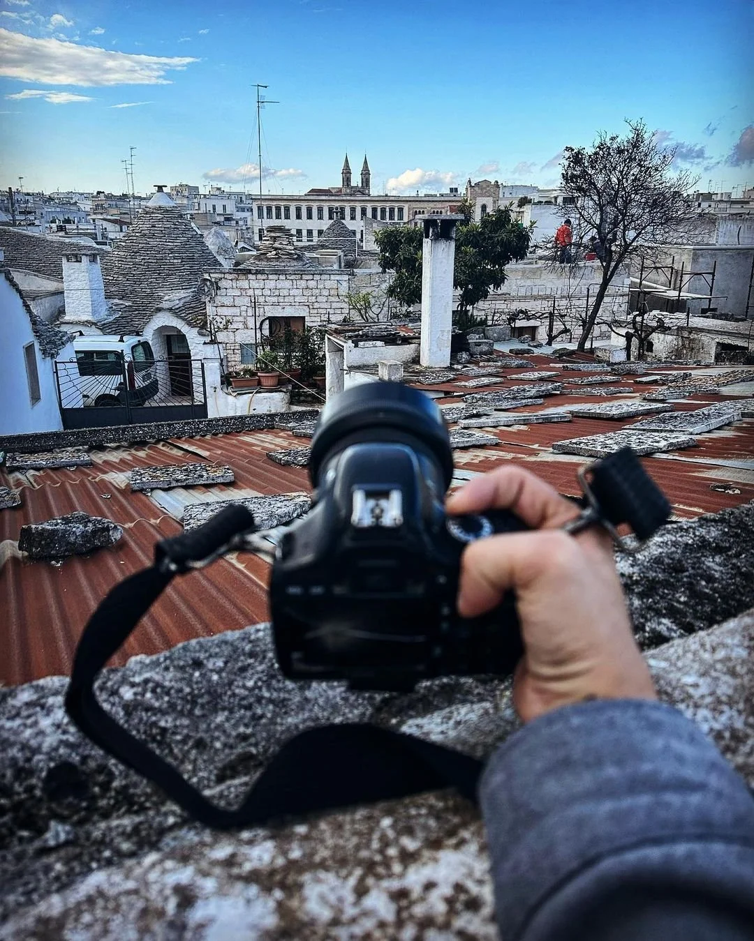 A person holding a camera on a rooftop, capturing a view of a European city with traditional white stone buildings, trees, and church steeples under a blue sky with clouds.