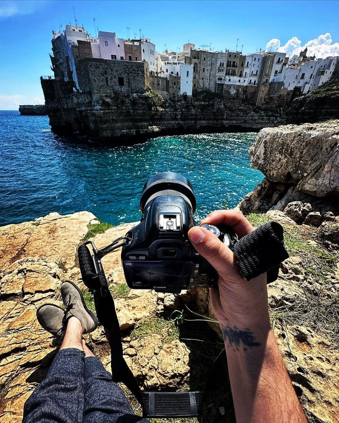 A person holding a camera on a rocky coastline taking a photo of the water and historic buildings on a cliff in the distance on a clear, sunny day.