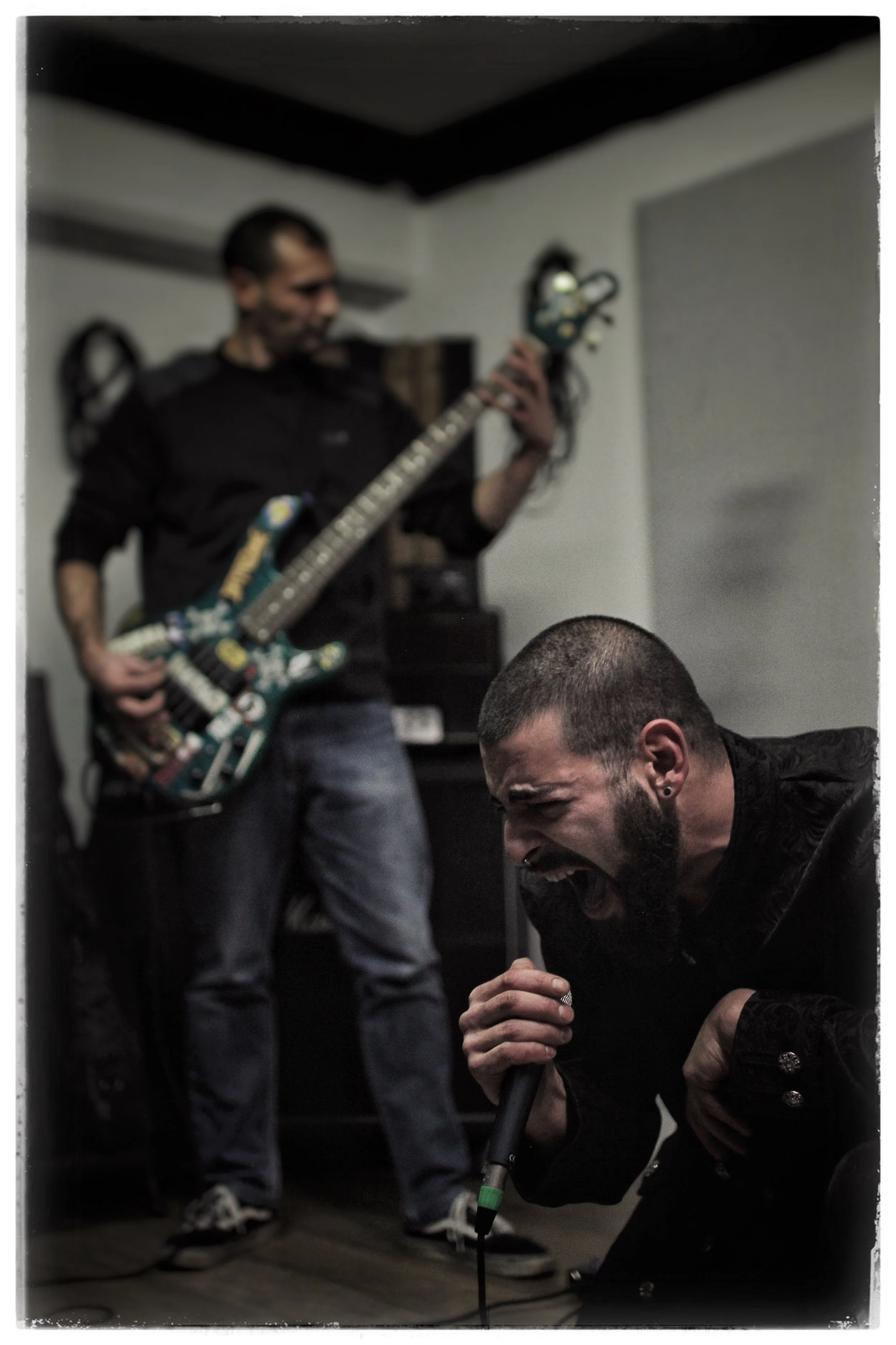 A man playing an electric guitar dressed in black stands in the background, while another man in a black jacket and jeans is kneeling in the foreground, singing into a microphone. The scene appears to be indoors, possibly during a practice or perform