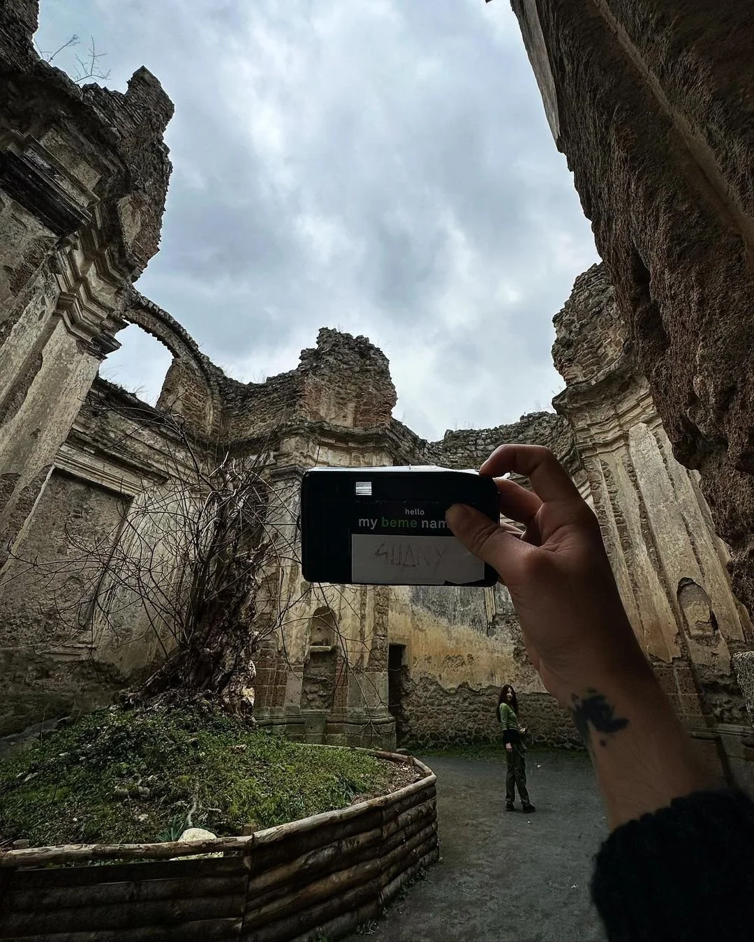 A person taking a photo of ancient ruins with a camera, standing inside the ruins which have weathered stone walls and an open sky overhead, with another person standing in the background.