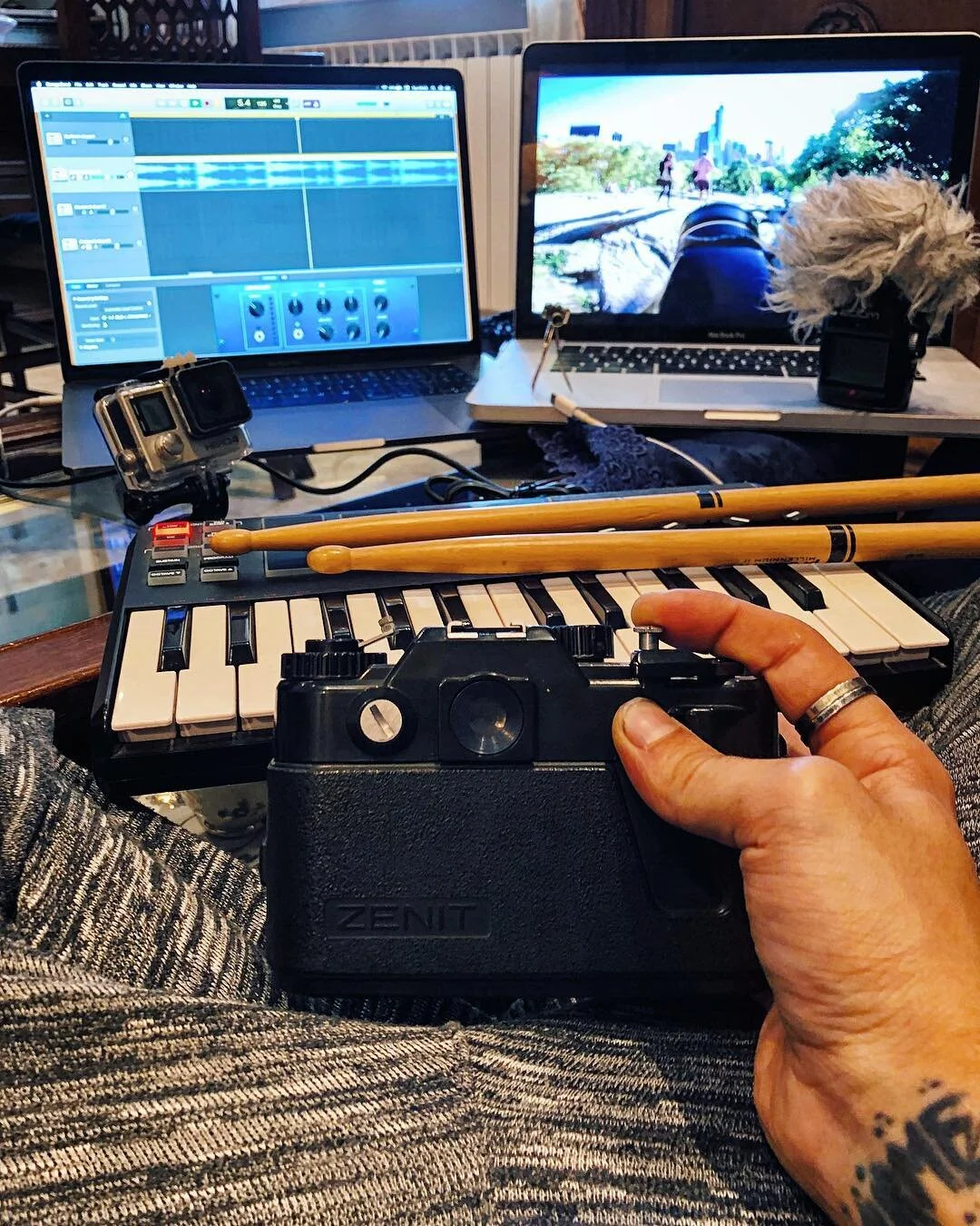 A person holding a Zenit camera with a keyboard and two drumsticks on the keyboard. In the background, there are two laptops, one displaying music editing software and the other showing a cityscape scene. An action camera is mounted on the keyboard w