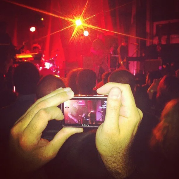 Person taking a photo of a live concert on a stage with a smartphone at a crowded indoor music venue.