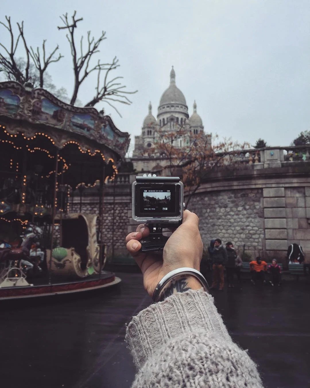 Person taking a photo of Sacré-Cœur Basilica in Paris with a camera, in a carnival-like setting with a carousel on the left and a group of people sitting and standing in front of a stone wall.