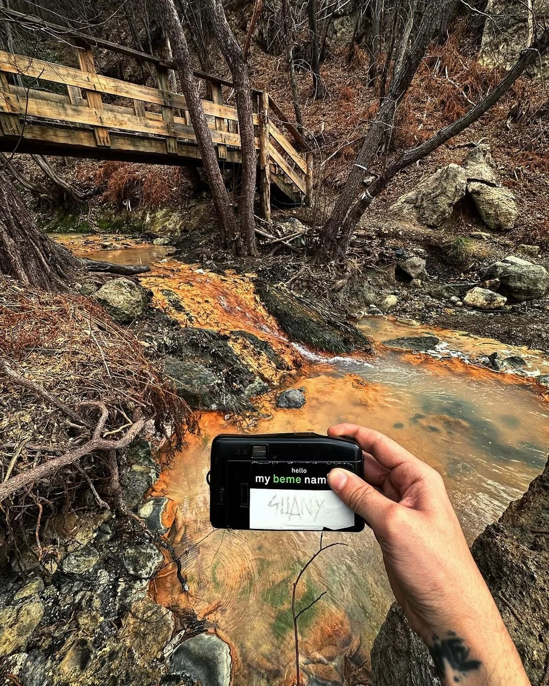 Person holding a camera with a note that says 'SHAY' and a partially visible message on the camera screen, in a natural landscape with a wooden bridge, trees, rocks, and a small stream with orange-colored water.